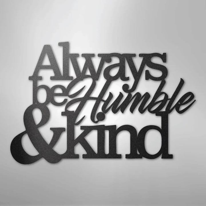 Black metal wall art with "Always be Humble & Kind" in varied fonts, casting a shadow on a white wall.