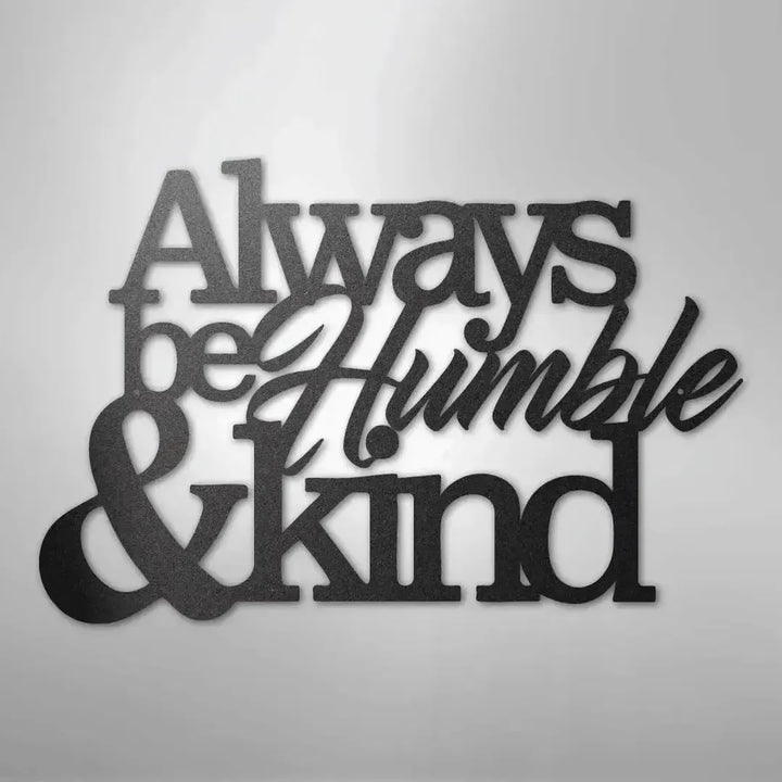 Black metal wall art with "Always be Humble & Kind" in varied fonts, casting a shadow on a white wall.