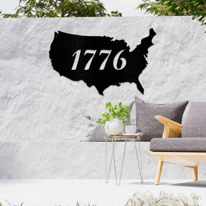 Black metal USA map wall art with "1776" cutout, displayed on a white outdoor wall above patio furniture.