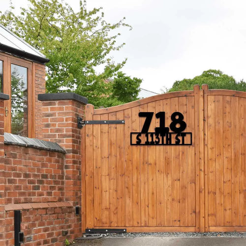 Black metal address sign "718 S 113TH ST" mounted on a rustic wooden gate, with brick wall beside it.
