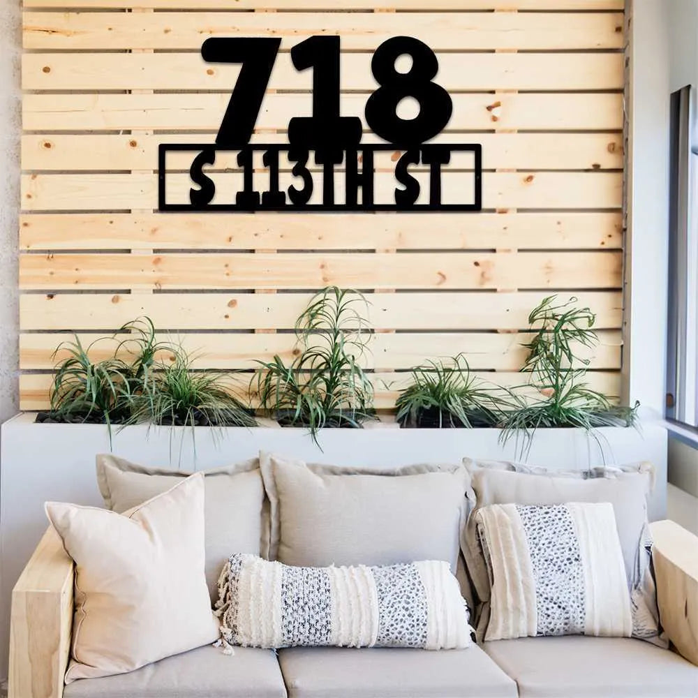 Black metal address sign "718 S 113TH ST" on a wooden slat wall above green plants and a sofa.