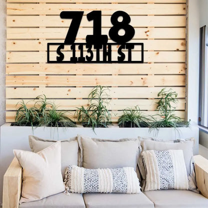 Black metal address sign "718 S 113TH ST" on a wooden slat wall above green plants and a sofa.