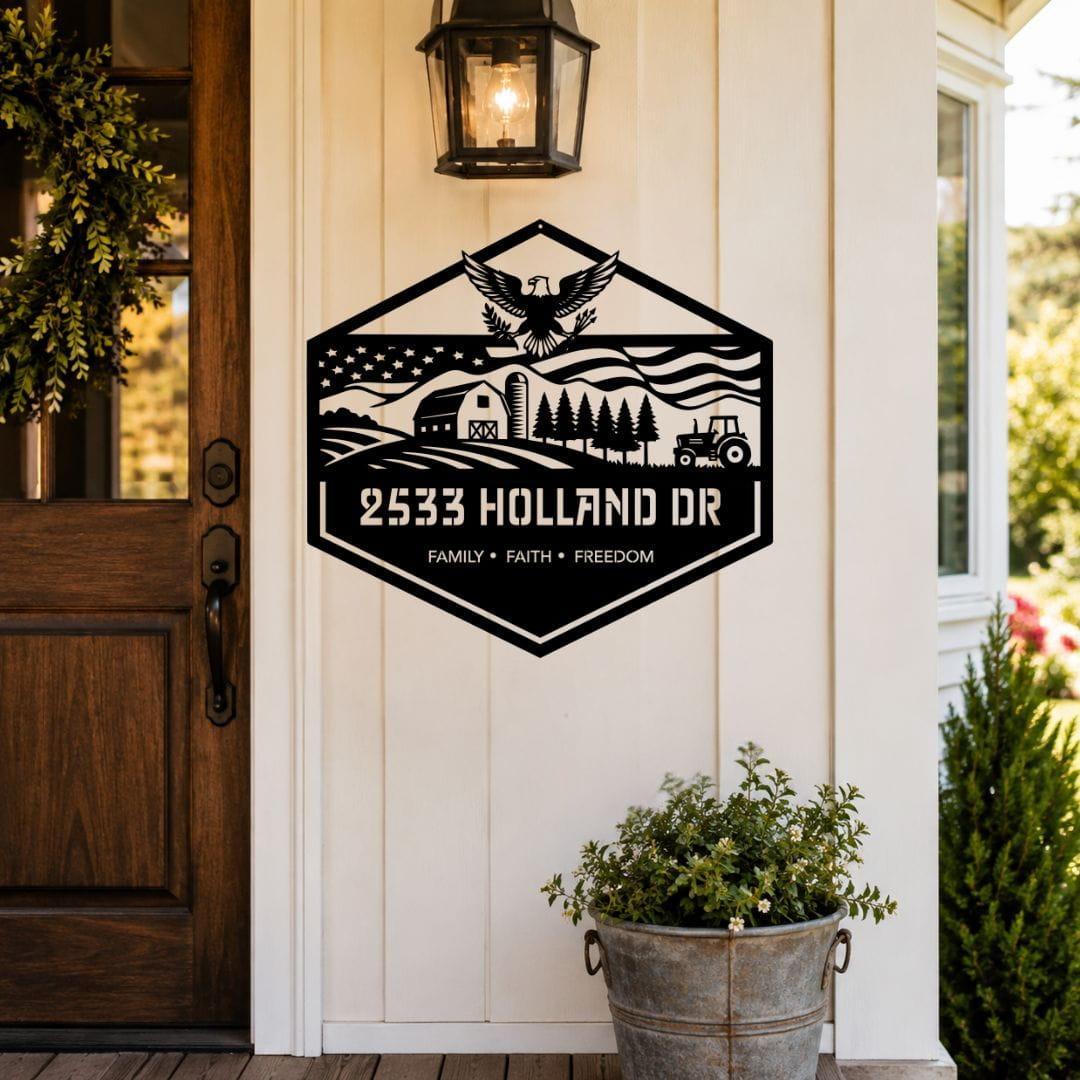A black metal farm-style sign mounted on a white porch wall, featuring an eagle, barn, trees, tractor, and the text '2533 HOLLAND DR' and 'FAMILY • FAITH • FREEDOM'. A wooden door, wall lantern, and potted plants are visible around it.