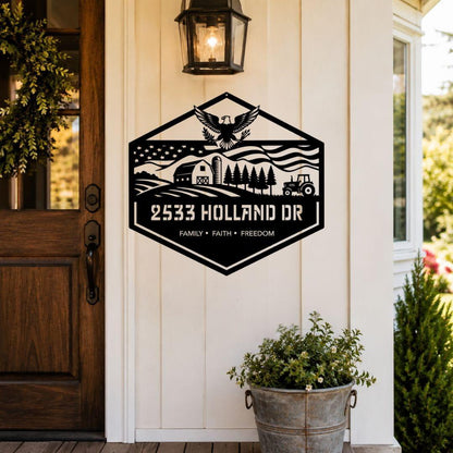 A black metal farm-style sign mounted on a white porch wall, featuring an eagle, barn, trees, tractor, and the text '2533 HOLLAND DR' and 'FAMILY • FAITH • FREEDOM'. A wooden door, wall lantern, and potted plants are visible around it.