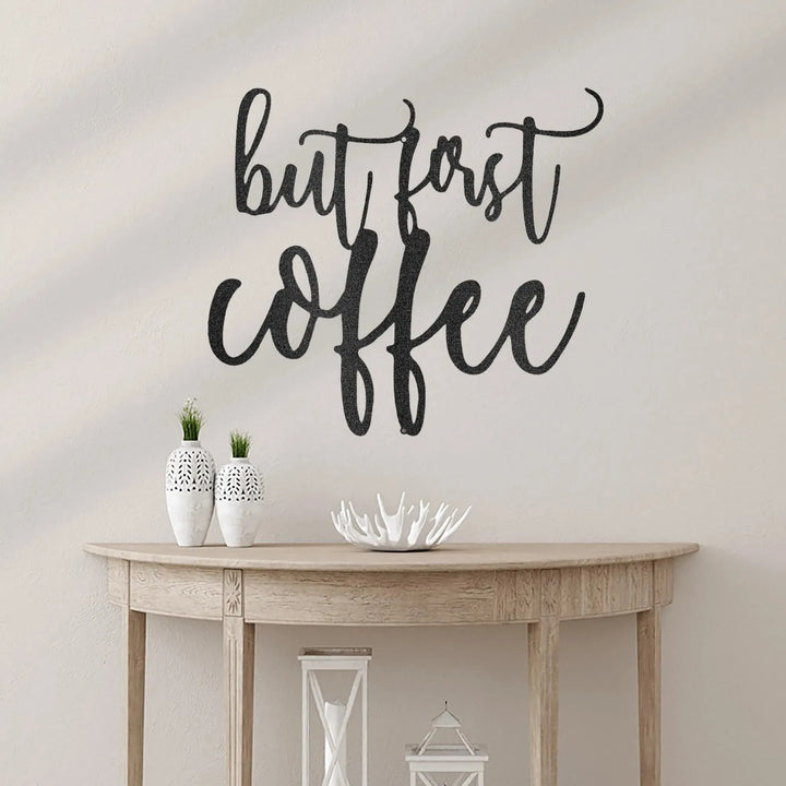 Black metal "but first coffee" script wall art above a light wood console table.
