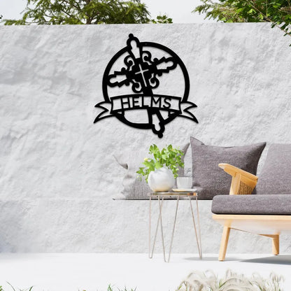 Black Catholic monogram metal wall art with a cross, fleur-de-lis, and "HELMS" banner, on a white outdoor wall.