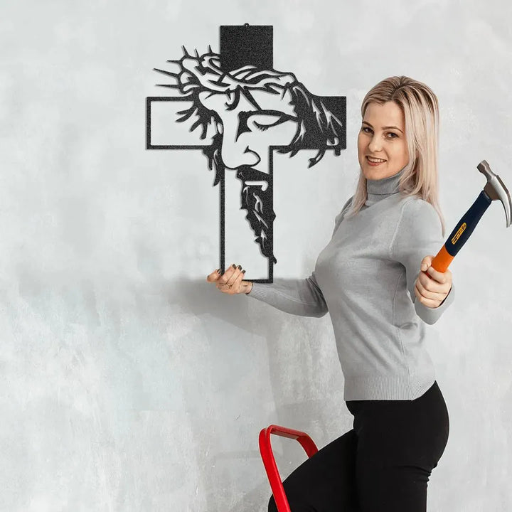 Black metal Christ cross wall art with Jesus' face, held by a woman on a ladder with a hammer.