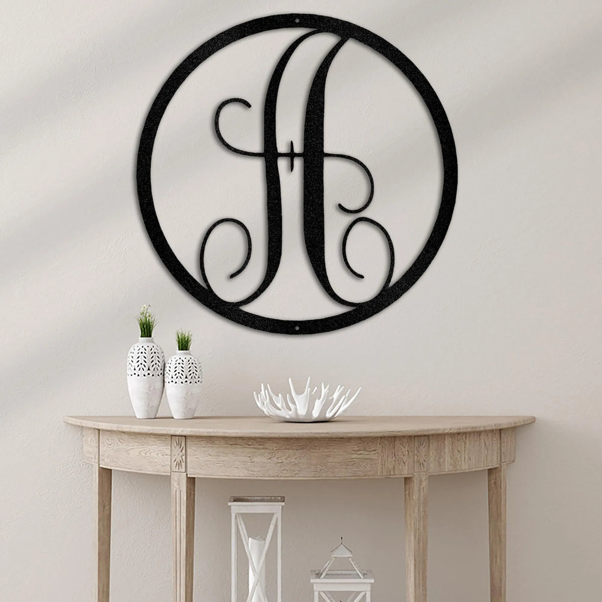 Black metal circle monogram wall art with a fancy script "H" on a light wall above a wooden console table.