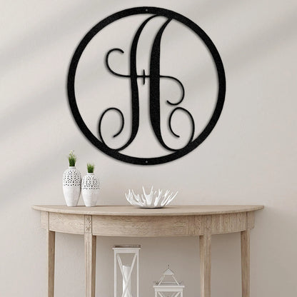 Black metal circle monogram wall art with a fancy script "H" on a light wall above a wooden console table.