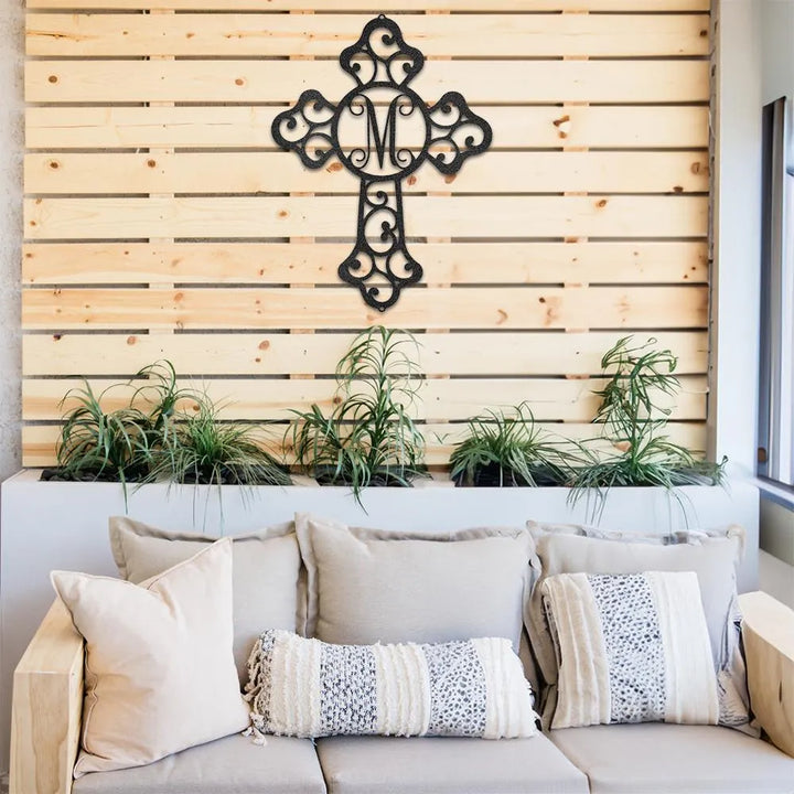 Black metal cross wall art with intricate scrollwork and a central "M" monogram, displayed on a wooden wall.