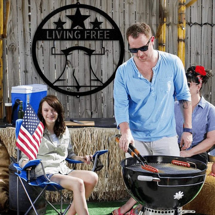 Black Liberty Bell "LIVING FREE" metal wall art above a family grilling outdoors.