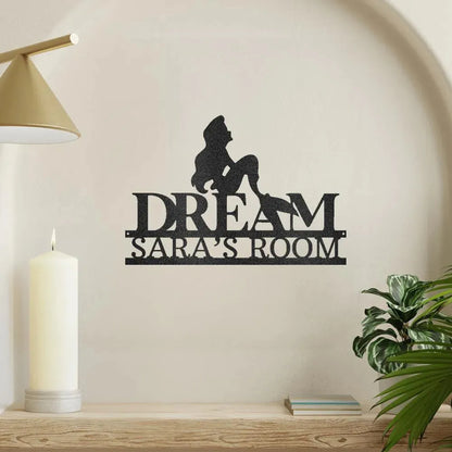 Black metal "DREAM SARA'S ROOM" wall art with a mermaid silhouette, on a shelf with a candle.