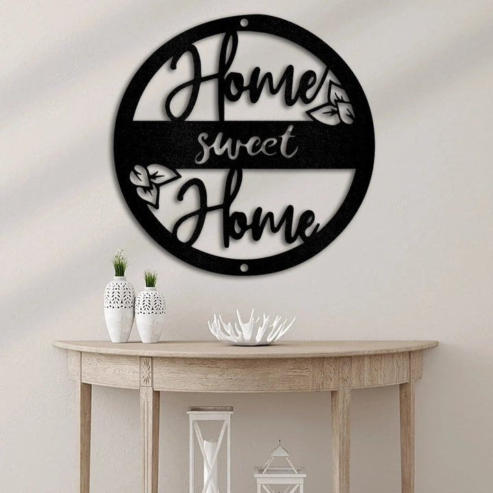 Black metal "Home Sweet Home" circle monogram wall art with leaf accents, displayed above a wooden console table.