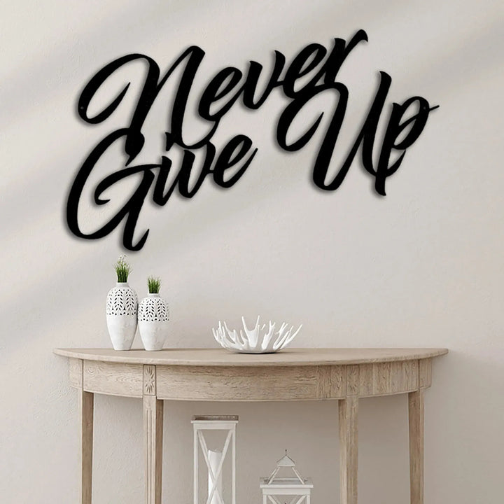 Black "Never Give Up" metal wall art in a script font, displayed above a light wood console table.