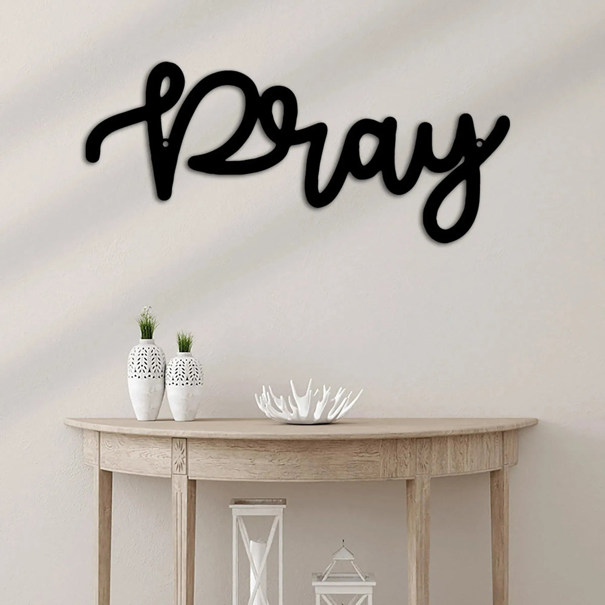 Black metal "Pray" script wall art in a flowing cursive font, mounted above a wooden console table.