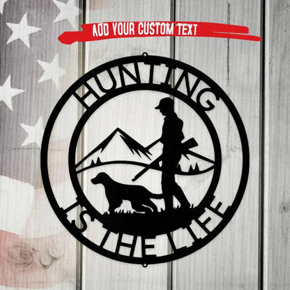 Black metal "Hunting Is The Life" wall art with hunter, dog, and mountains, on a rustic wood background.