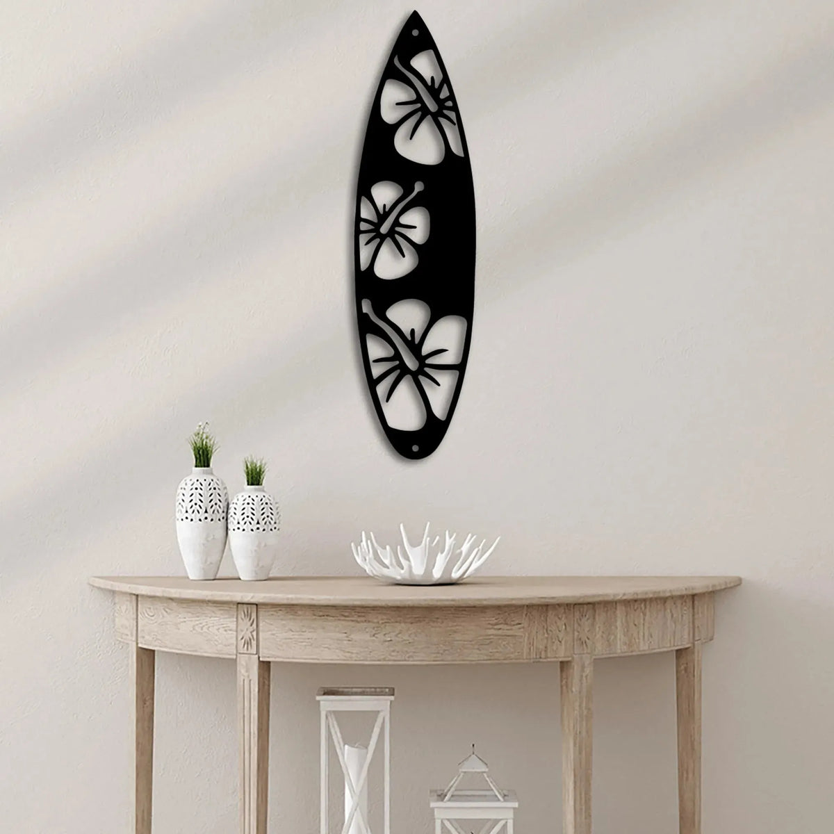 Black metal surfboard wall art with three white hibiscus flower cutouts, hanging above a wooden console table.