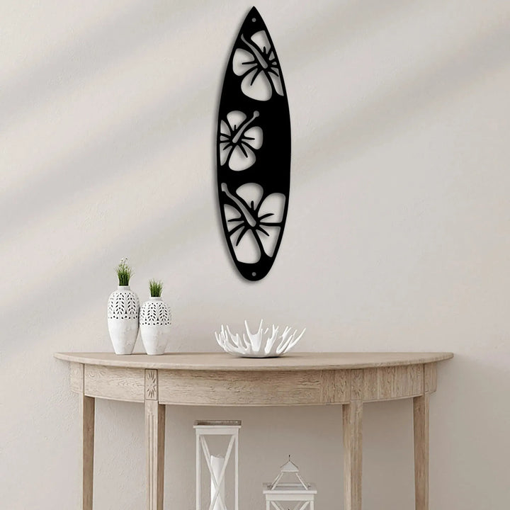 Black metal surfboard wall art with three white hibiscus flower cutouts, hanging above a wooden console table.
