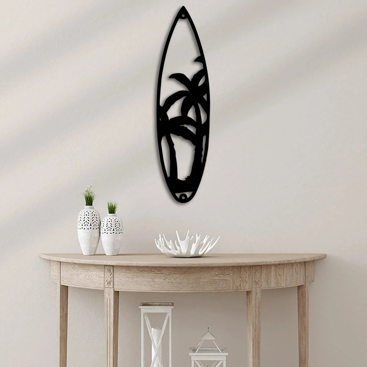Black metal wall art shaped like a surfboard with palm trees, hanging above a light wood console table.