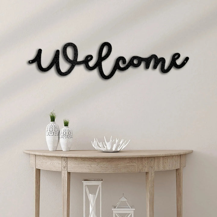 Black script "Welcome" metal wall art above a light wood console table with white vases.