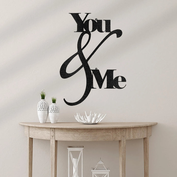 Black metal "You & Me" script wall art on a light wall above a wooden console table.