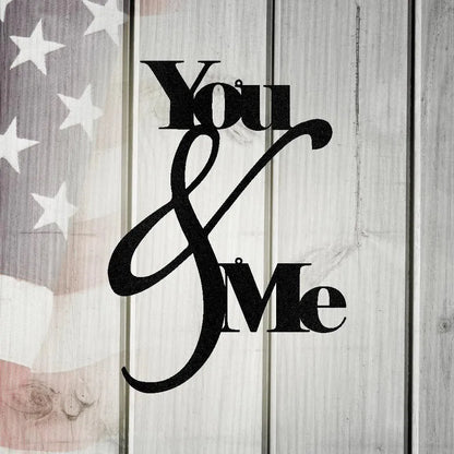Black metal "You & Me" quote wall art on a rustic white wooden background with a faint American flag.